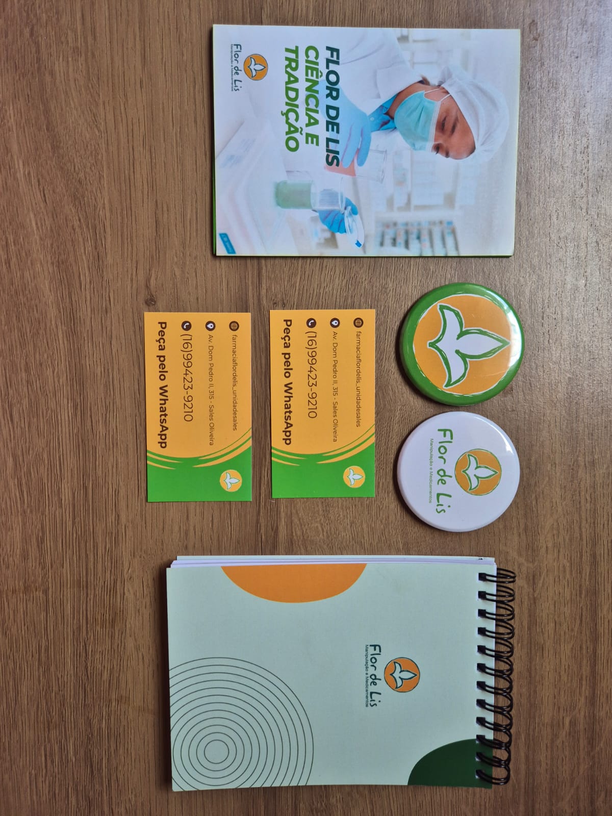 Materiais gráficos da farmácia Flor de Lis. Folder A6 com 3 dobras com o título Ciência e Tradição, bottoms com o logotipo da farmácia, cartões de visitas com dados de contato da farmácia e bloco de anotações personalizado com logotipo da farmácia.